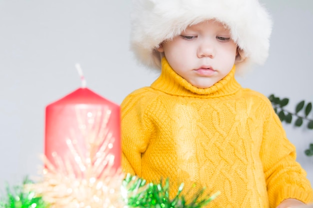 Un mignon petit garçon dans un pull jaune et un chapeau de Père Noël rouge avec une bougie de Noël rouge