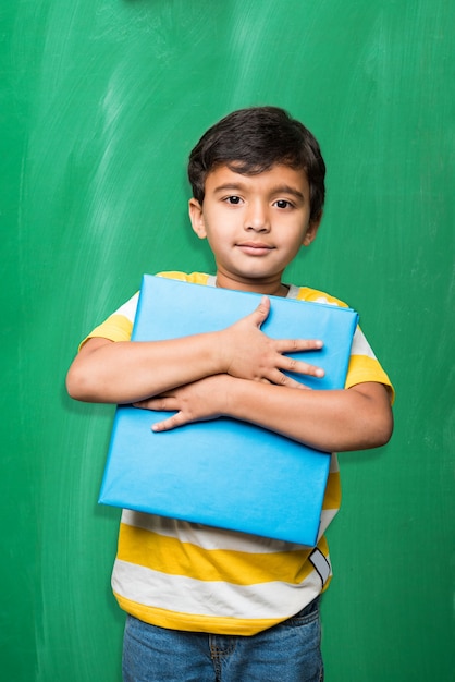 Mignon petit garçon de l'école indienne à la main pose étirée sur fond de tableau vert ou de tableau de craie tenant des livres, coupe de la victoire, etc., isolé