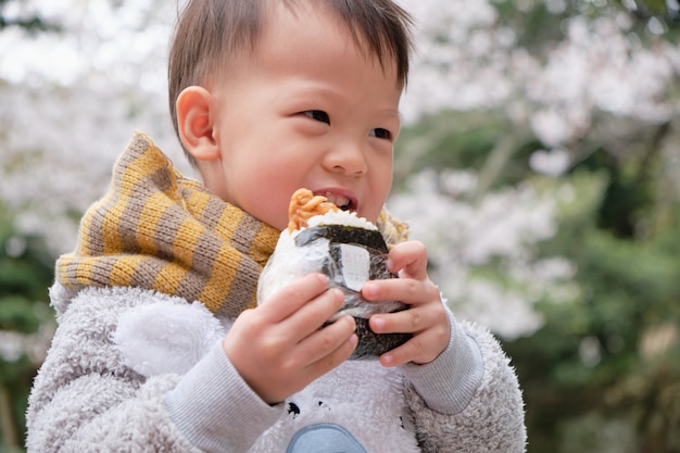 Mignon petit garçon enfant en bas âge asiatique mordre et manger Onigiri, nourriture japonaise, boule de riz japonaise, triangle de riz aux algues dans le jardin de printemps en fleurs tout en visitant sakura