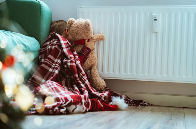 Mignon petit garçon enveloppé d'un plaid d'identification assis près d'un radiateur chauffant les pattes d'ours en peluche