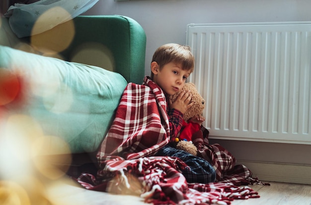 Mignon petit garçon enveloppé d'un plaid d'identification assis près d'un radiateur