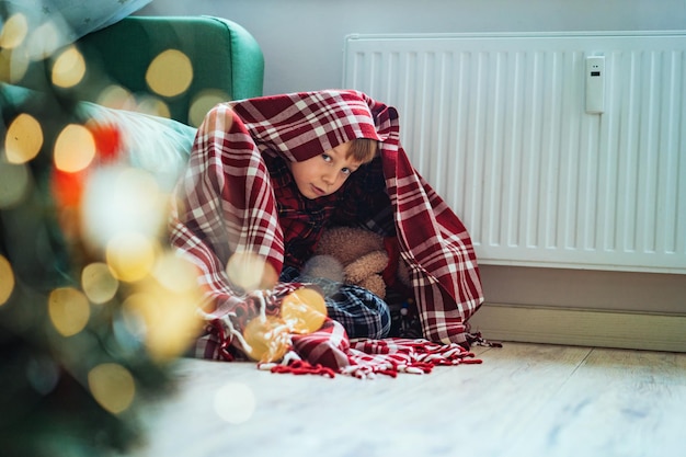 Mignon petit garçon enveloppé d'un plaid d'identification assis près d'un radiateur