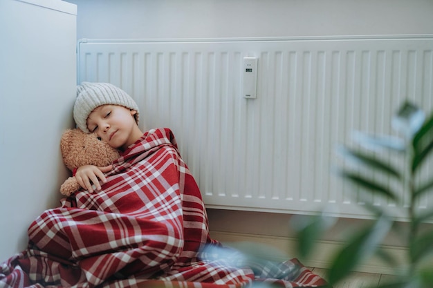 Mignon petit garçon enveloppé d'un plaid d'identification portant un bonnet en tricot assis près d'un radiateur étreignant un ours en peluche froid à la maison