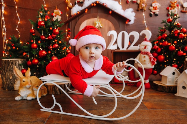 Mignon petit garçon habillé en père Noël sur un vieux traîneau vintage avec des cadeaux près de l'arbre de Noël, concept de joyeuses fêtes. 2022