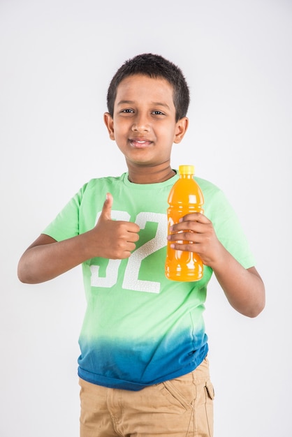 Mignon petit garçon indien ou asiatique avec une bouteille pour animaux de compagnie de boisson froide à l'orange ou à la mangue ou de jus de fruits, buvant ou tenant debout isolé sur fond bleu ou blanc.