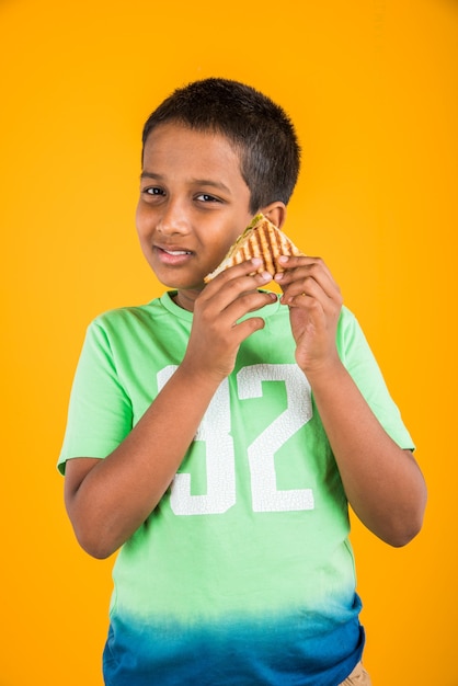 Mignon petit garçon indien ou asiatique mangeant de savoureux hamburgers, sandwichs ou pizzas dans une assiette ou une boîte. Debout isolé sur fond bleu ou jaune.