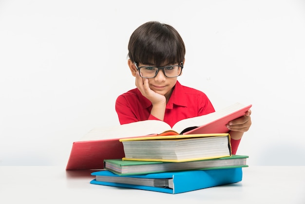 Mignon petit garçon indien ou asiatique tenant ou lisant un livre sur une table d'étude ou sur un sol blanc, isolé sur fond blanc