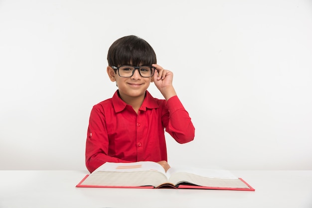 Mignon petit garçon indien ou asiatique tenant ou lisant un livre sur une table d'étude ou sur un sol blanc, isolé sur fond blanc