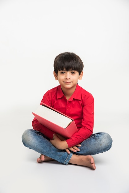 Mignon petit garçon indien ou asiatique tenant ou lisant un livre sur une table d'étude ou sur un sol blanc, isolé sur fond blanc