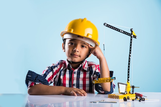 Mignon petit garçon indien jouant avec une grue jouet portant un chapeau de construction jaune ou un casque, concept d'enfance et d'éducation, isolé sur un tableau vert