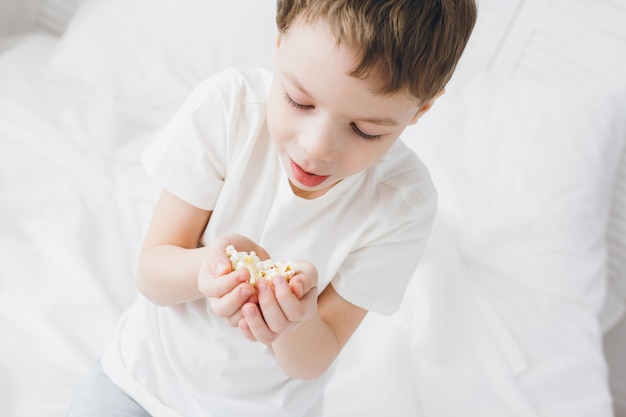 Mignon petit garçon mangeant du pop-corn assis dans son lit avec des draps blancs