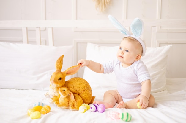 Mignon petit garçon avec des oreilles de lapin et des oeufs colorés sur un lit blanc à la maison jouant petit bébé blond avec un lapin concept de joyeuses pâques