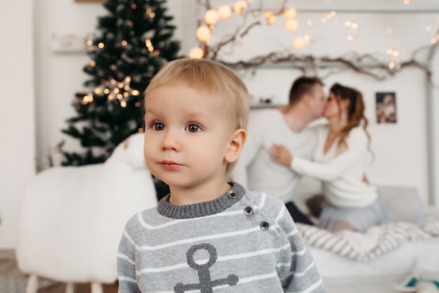 Mignon petit garçon en pull gris debout en studio et à la recherche de suite tandis que les jeunes parents amoureux étreignant et s'embrassant derrière