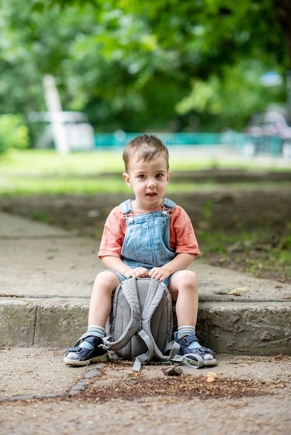 Un mignon petit garçon regarde la caméra tient une sacoche dans ses mains Vêtu d'une salopette en jean Earl