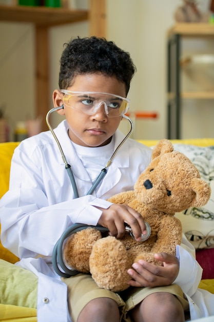 Mignon petit garçon réussi d'ethnie africaine en blanchon ayant des lunettes médicales de protection sur la tête et un stéthoscope sur le cou