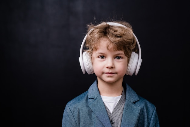 Mignon petit garçon en tenue décontractée, écouter de la musique dans un casque blanc devant la caméra sur un espace noir