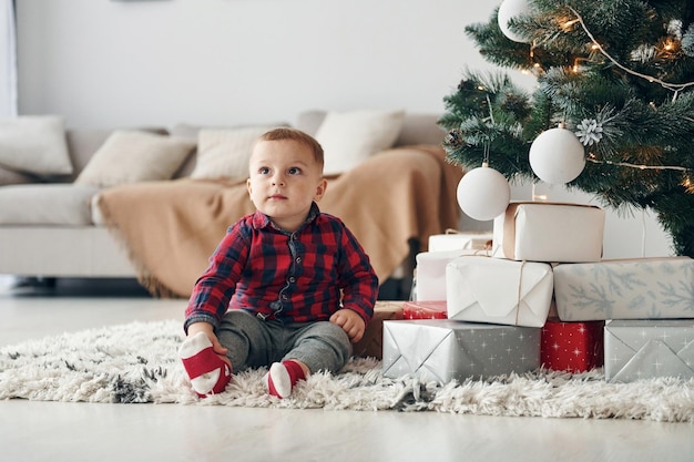 Mignon petit garçon en vêtements de fête est à l'intérieur près de l'arbre de noël au moment du nouvel an