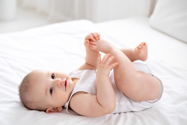 un mignon petit garçon vêtu d'un body blanc est allongé sur le dos sur un lit sur une literie blanche dans la chambre jouant avec ses bras et ses jambes portrait d'un enfant mignon aux yeux bruns et aux cheveux blonds