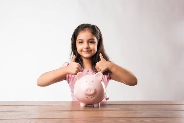 Mignon petit indien ou asiatique avec tirelire, mettant des pièces de monnaie, ou pensant ou montrant des pouces vers le haut. Planifier pour l'avenir