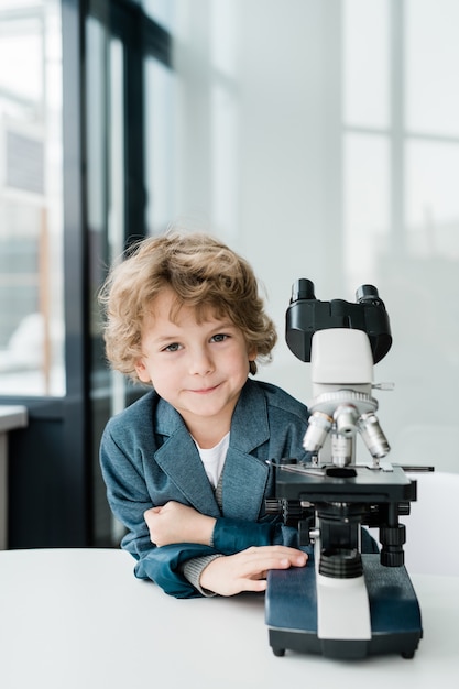 Mignon petit scientifique qui réussit vous regarde en se tenant debout au microscope en classe ou en laboratoire