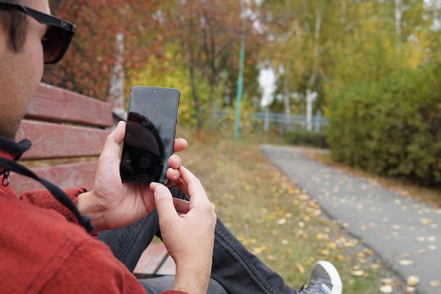 Mignon Séduisant Beau Mec Sérieux Portant Des Vêtements Décontractés Et Des Lunettes De Soleil à L'extérieur Tenant Un Smartphone Lisant Des Sms Travailler Via Un Smartphone Correspondance Sur Un Réseau Social étudiant Dans Le Parc