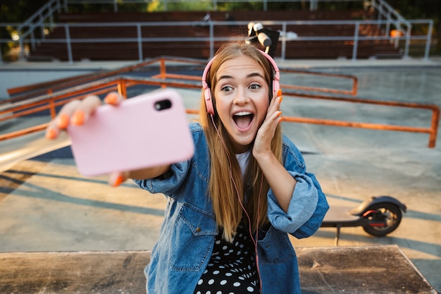 mignon surpris choqué positif jeune adolescente à l'extérieur dans le parc en écoutant de la musique avec des écouteurs tenant un téléphone portable prendre un selfie.