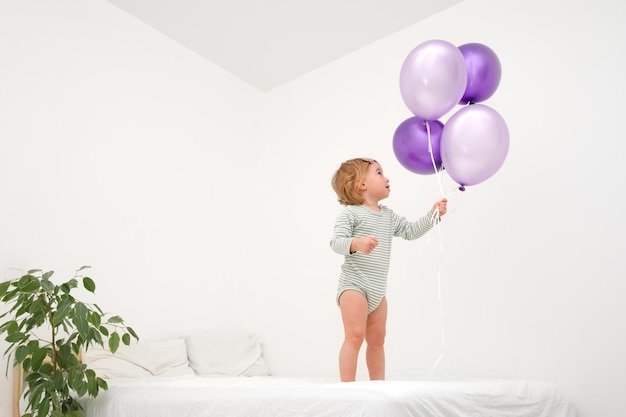 Mignonne drôle de bébé blonde caucasienne tout-petit de 12 ans jouant sur le lit avec de gros ballons violets de couleur très tendance péri sur fond blanc dans un intérieur scandinave moderne à la maisonCopier l'espace