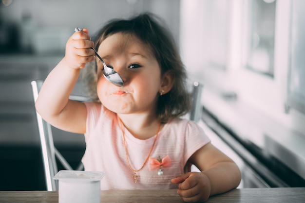 Mignonne, drôle de petite fille mangeant du yaourt dans la cuisine dans une robe rose dans l'après-midi mignon
