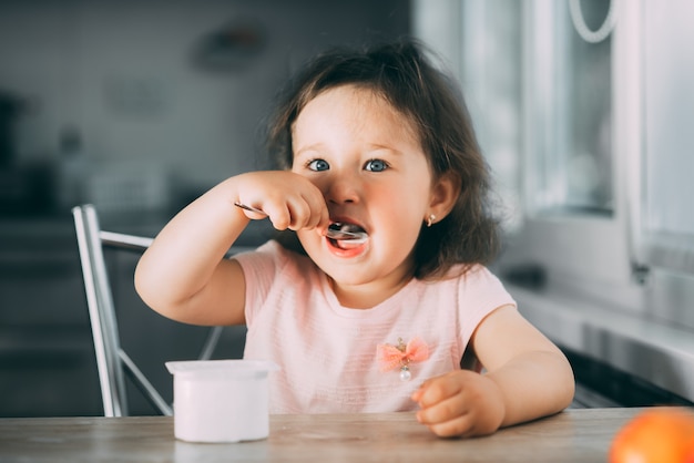 Mignonne, drôle de petite fille mangeant du yaourt dans la cuisine dans une robe rose dans l'après-midi mignon