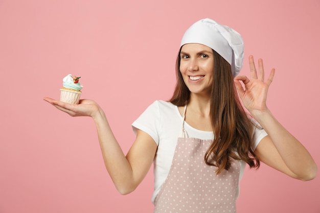 Mignonne femme au foyer chef cuisinier pâtissier boulanger en t-shirt blanc, chapeau de chefs toque cuisson tenir doux isolé sur fond pastel rose en studio. Processus de fabrication de cupcakes. Maquette du concept de nourriture de l'espace de copie.