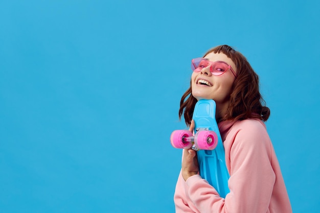 Mignonne jolie dame rousse souriante en lunettes de soleil à capuche rose avec penny board regarde la caméra posant isolé sur fond de studio bleu Copie espace bannière offre mode cinéma activité de vacances