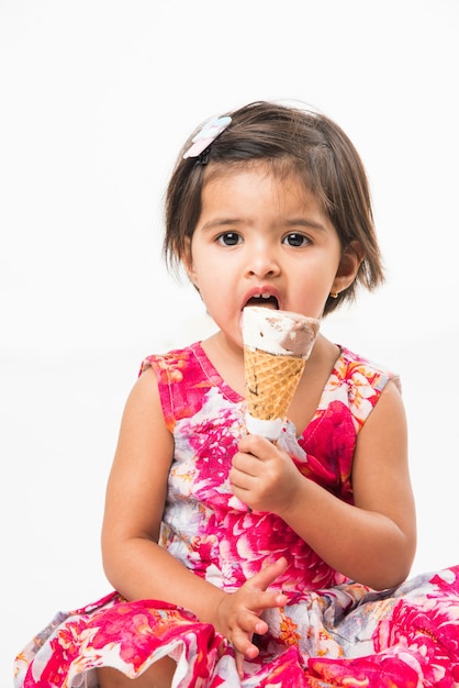 Mignonne petite fille asiatique indienne léchant ou mangeant de la crème glacée au chocolat en cône, isolée sur fond blanc