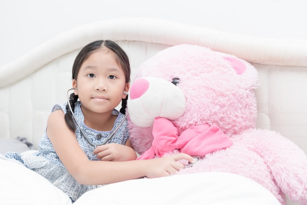 mignonne petite fille asiatique jouant stéthoscope avec gros ours en peluche rose sur le lit, concept heureux