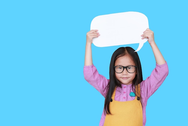 Mignonne petite fille asiatique tenant une bulle de dialogue vide vide pour dire quelque chose en souriant et en regardant directement la caméra isolée sur fond bleu Concept d'éducation et de conversation