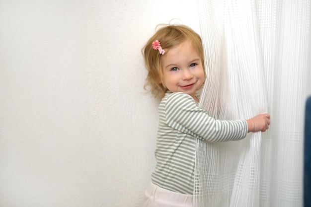 Mignonne petite fille blonde caucasienne de 12 ans jouant avec des rideaux blancs s'amusant à la maison dans un intérieur lumineux regardant la caméra