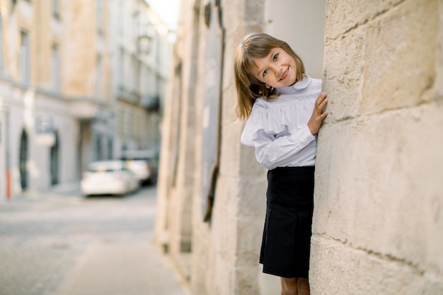 Mignonne petite fille blonde caucasienne en vêtements noir et blanc, posant devant la caméra à l'extérieur dans la vieille rue de la ville, cachant son visage derrière l'ancien mur du bâtiment