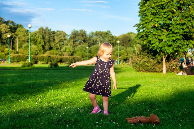 Mignonne petite fille blonde de deux ans jouant avec un ours en peluche sur une herbe verte fraîche avec des fleurs. Enfant s'amusant à faire ses premiers pas sur une pelouse naturelle tondue. Concept d'enfance heureuse et saine