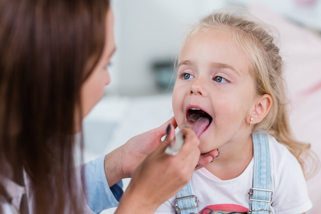 Mignonne petite fille blonde ouvrant la bouche alors qu'il était assis devant un clinicien examinant sa gorge avec une spatule en stell