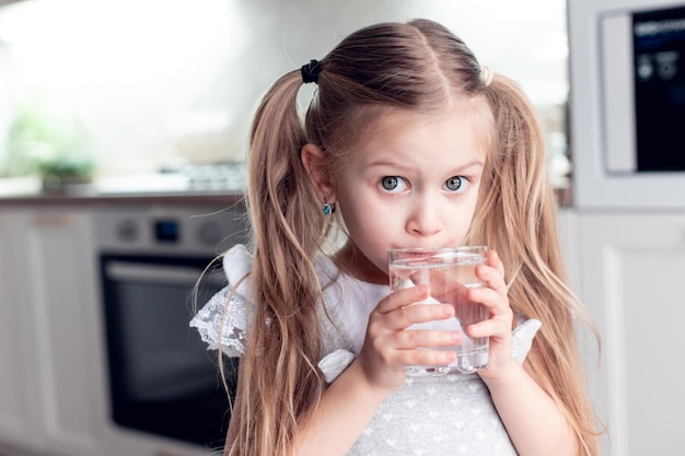 Mignonne petite fille boit de l'eau douce propre à partir d'un verre transparent à la maison dans la cuisine pro utile
