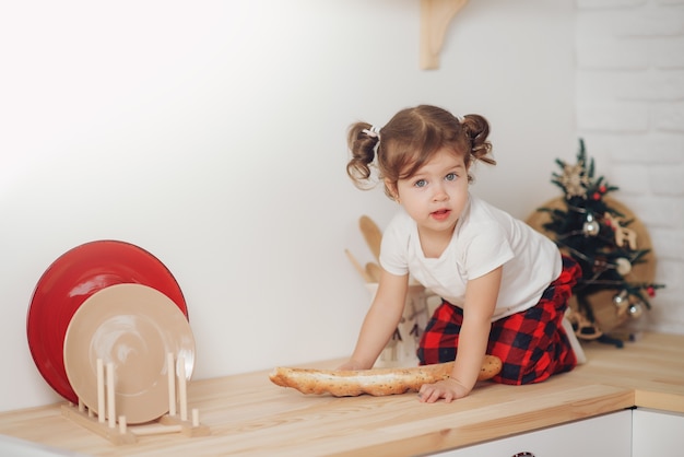 Mignonne petite fille en bonnet de Noel, préparer des biscuits dans la cuisine à la maison. S'assoit sur la table de la cuisine et aide maman à préparer un dîner de Noël festif