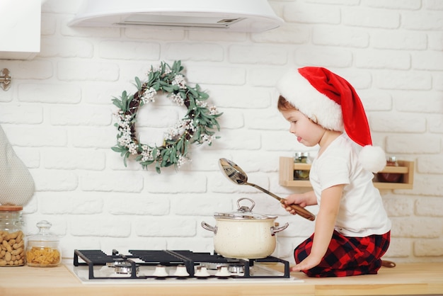 Mignonne petite fille en bonnet de Noel, préparer des biscuits dans la cuisine à la maison. S'assoit sur la table de la cuisine et aide maman à préparer un dîner de Noël festif