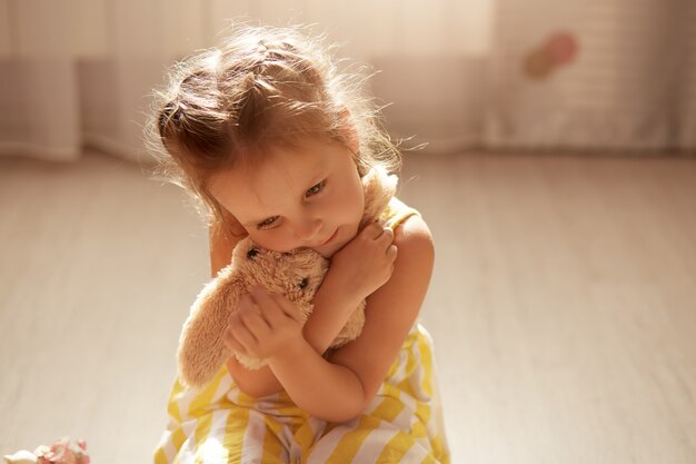 Mignonne Petite Fille Bouleversée Vêtue D'une Robe Jaune Et Blanche Est Assise Sur Le Sol Avec Sa Peluche Dans Les Mains Et La Serre Dans Ses Bras, Enfant De Sexe Féminin Assis Sur Le Sol.