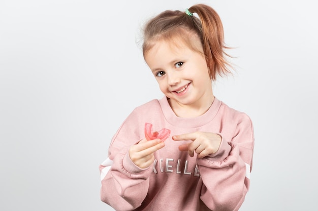 Mignonne petite fille caucasienne aux cheveux blonds tient un entraîneur myofonctionnel dentaire rose sur fond blanc. le tariner dentaire est conçu pour aider à égaliser la croissance des dents et à corriger la morsure