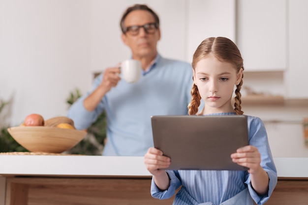 Mignonne petite fille concentrée assis à la maison et jouer à des jeux sur tablette pendant que grand-père se repose