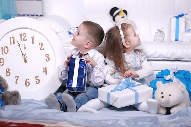 Mignonne petite fille et garçon avec des coffrets cadeaux et grande horloge blanche assis près du canapé blanc à Noël et nouvel an