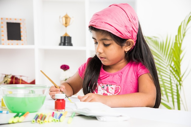 Mignonne petite fille indienne ou asiatique appréciant le dessin OU la peinture avec un pinceau et de la peinture sur papier à la maison