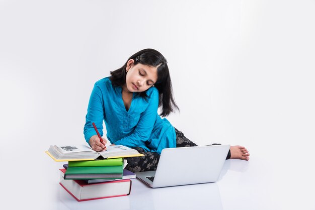 Mignonne petite fille indienne ou asiatique étudiant sur un ordinateur portable ou travaillant sur un projet scolaire en position couchée ou assise sur le sol, isolée sur fond blanc