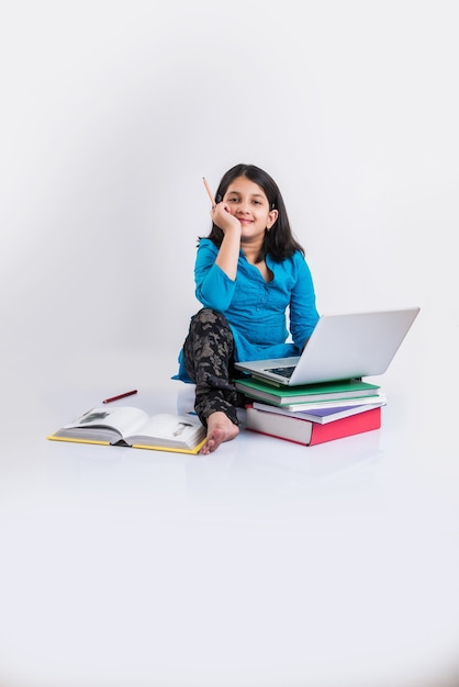 Mignonne petite fille indienne ou asiatique étudiant sur un ordinateur portable ou travaillant sur un projet scolaire en position couchée ou assise sur le sol, isolée sur fond blanc