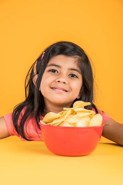 Mignonne petite fille indienne ou asiatique mangeant des chips ou des gaufrettes de pommes de terre dans un grand bol rouge, sur fond jaune