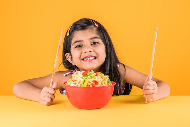 Mignonne petite fille indienne ou asiatique mangeant de délicieuses nouilles chinoises avec une fourchette ou des baguettes, isolée sur fond coloré